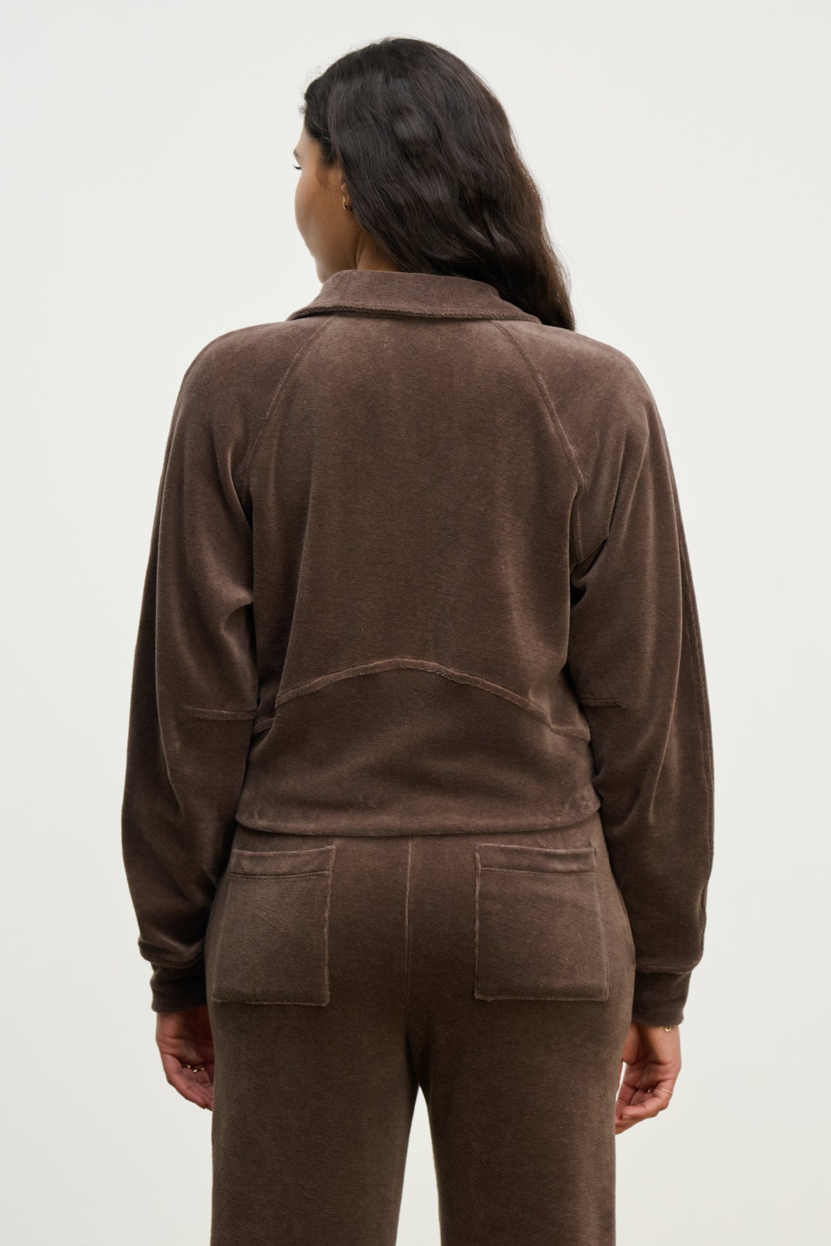 A woman with long dark hair is seen from the back, wearing the CLAIRE HOODIE by Velvet by Graham & Spencer in brown cotton velour with matching pants and patch pockets, standing against a plain light background.-39814826754241