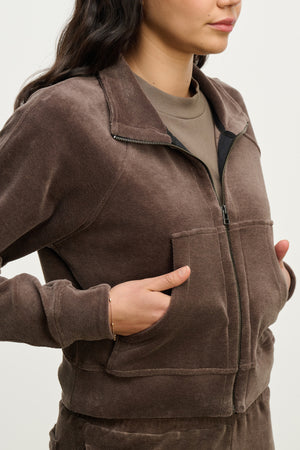 Wearing the CLAIRE JACKET by Velvet by Graham & Spencer—a brown zip-up cotton velour hoodie with large front pockets, shown from shoulders to hips, hands in pockets—makes for a cozy staple in any casual wardrobe.