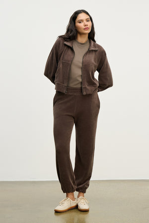 A woman stands against a plain background wearing the ESSIE SWEATPANT by Velvet by Graham & Spencer, a brown cotton velour jogger with side seam pockets, paired with a matching jacket, taupe t-shirt, and beige sneakers.