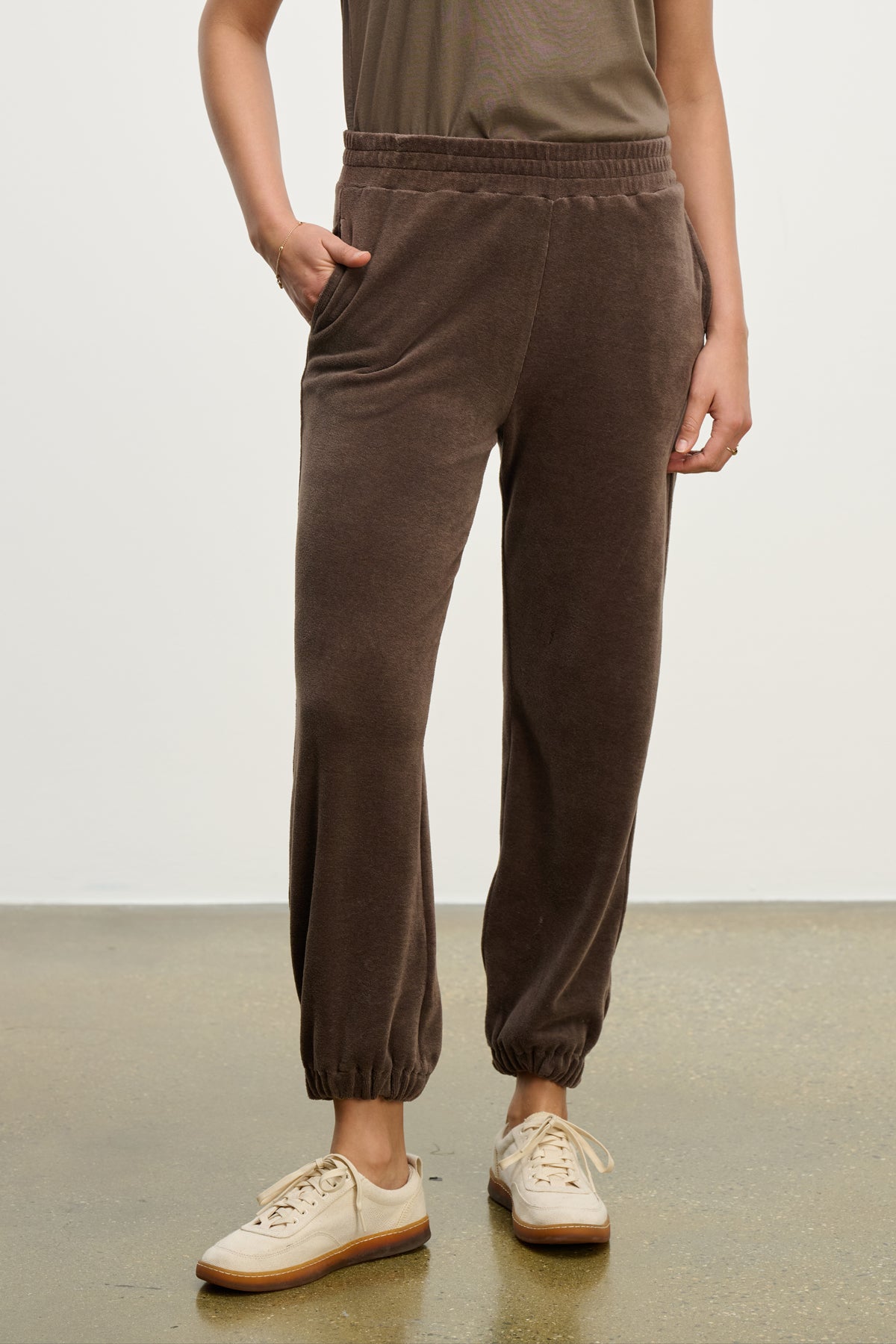   Someone wearing the ESSIE SWEATPANT by Velvet by Graham & Spencer, featuring brown cotton velour and side seam pockets, pairs them with a taupe shirt and beige sneakers, posing with one hand in a pocket against a plain background. 