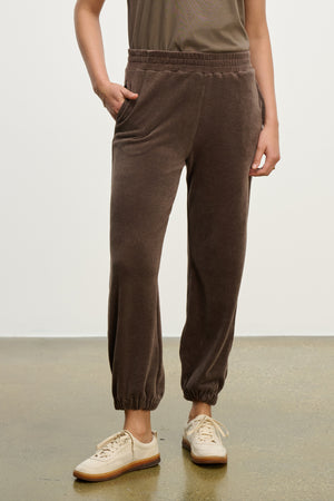 Someone wearing the ESSIE SWEATPANT by Velvet by Graham & Spencer, featuring brown cotton velour and side seam pockets, pairs them with a taupe shirt and beige sneakers, posing with one hand in a pocket against a plain background.
