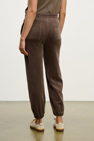 A person stands on a smooth floor, facing away from the camera, wearing Velvet by Graham & Spencer's ESSIE SWEATPANT with back and side seam pockets, paired with beige sneakers.