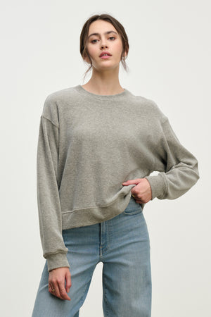 A woman stands against a plain light background in the HALIMA PULLOVER by Velvet by Graham & Spencer—a loose, grey cotton velour crewneck—paired with light blue jeans for an effortlessly stylish loungewear look.