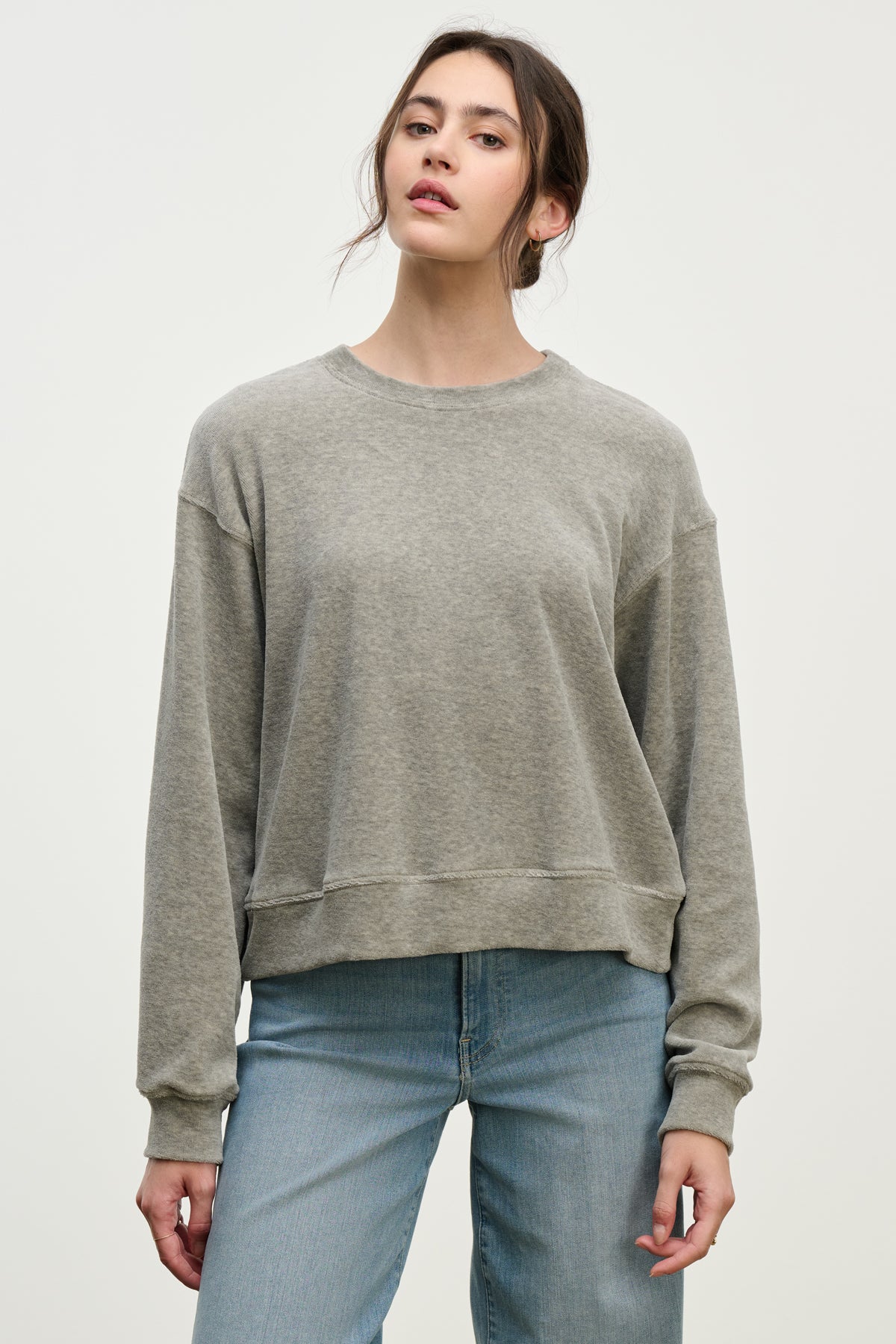 A woman stands against a plain background in the HALIMA PULLOVER by Velvet by Graham & Spencer, a loose-fit gray cotton velour top with a crew neckline, paired with light blue jeans and her hair pulled back for a cozy loungewear vibe.