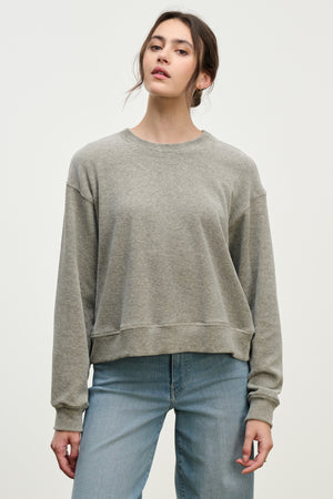 A woman stands against a plain background in the HALIMA PULLOVER by Velvet by Graham & Spencer, a loose-fit gray cotton velour top with a crew neckline, paired with light blue jeans and her hair pulled back for a cozy loungewear vibe.