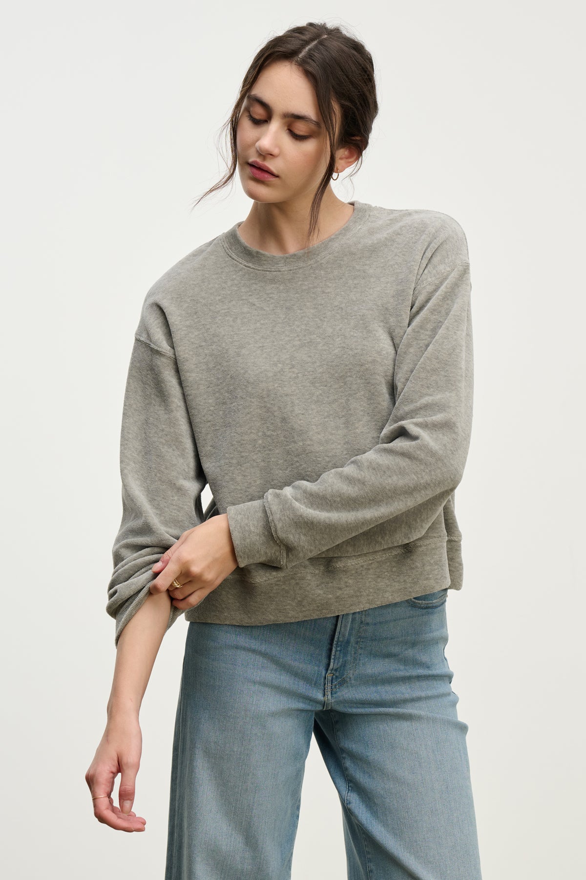 Wearing the HALIMA PULLOVER by Velvet by Graham & Spencer and light blue jeans, a person rolls up their sleeve and looks down while standing against a plain background.-Hover Image