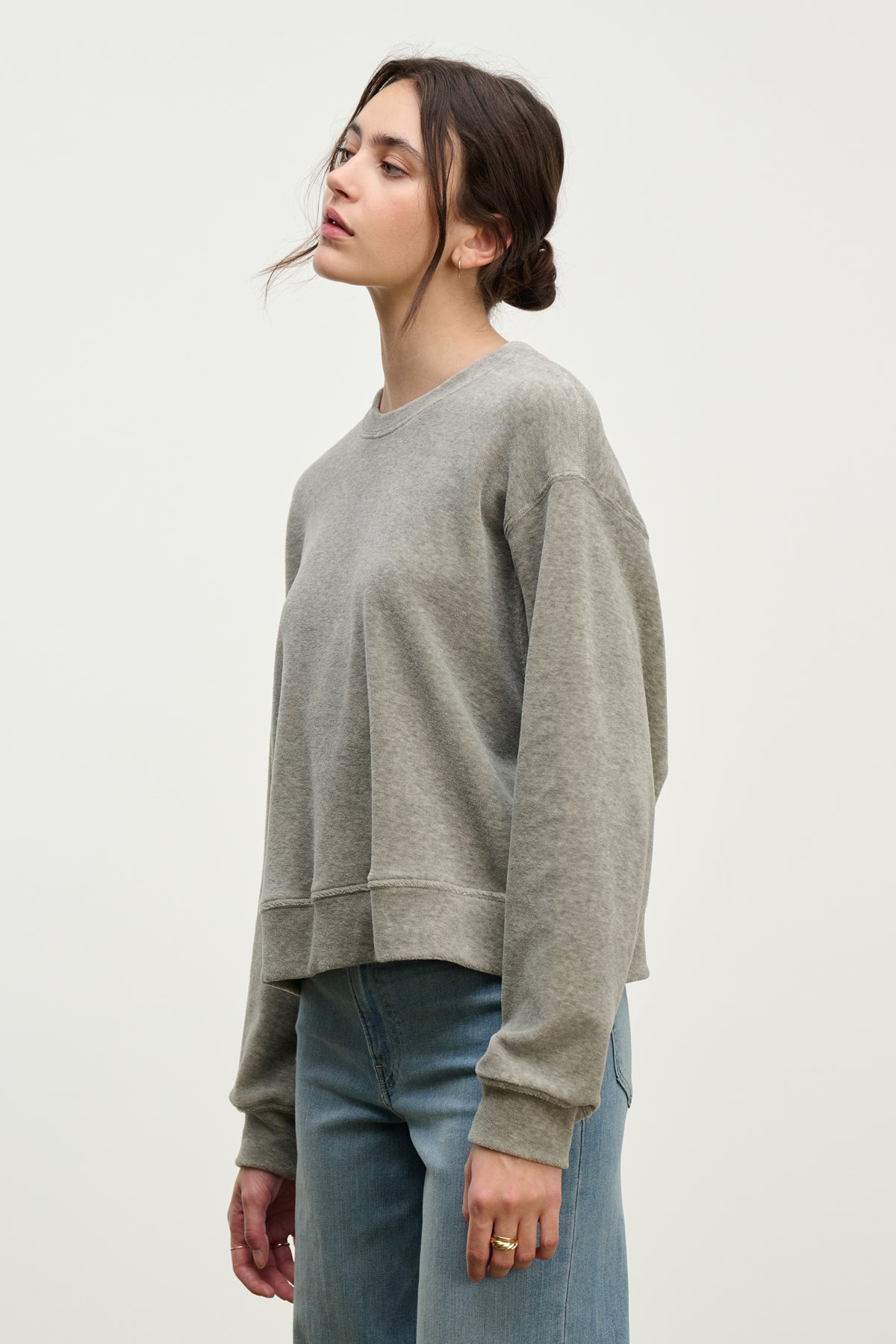 A person with dark hair in a low bun models the HALIMA PULLOVER by Velvet by Graham & Spencer—a loose grey cotton velour crewneck—paired with blue jeans, standing against a plain light background.-39800325308609