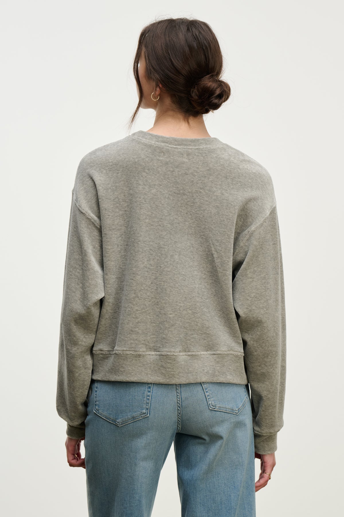 A woman with brown hair in a low bun wears the HALIMA PULLOVER by Velvet by Graham & Spencer in gray cotton velour with a crew neckline and blue jeans, standing back to the camera against a light background—effortless loungewear style.-39800325275841