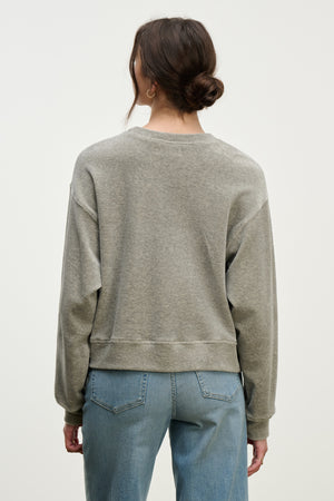 A woman with brown hair in a low bun wears the HALIMA PULLOVER by Velvet by Graham & Spencer in gray cotton velour with a crew neckline and blue jeans, standing back to the camera against a light background—effortless loungewear style.