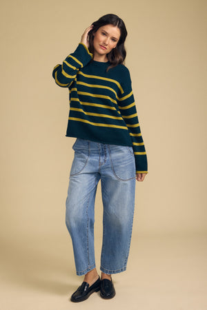Dressed against a beige background, the person wears Velvet by Graham & Spencer’s LUCIA SWEATER—a relaxed-fit dark green and yellow striped knit with a timeless silhouette—paired with loose-fitting blue jeans and black loafers, their hand gently touching their hair.