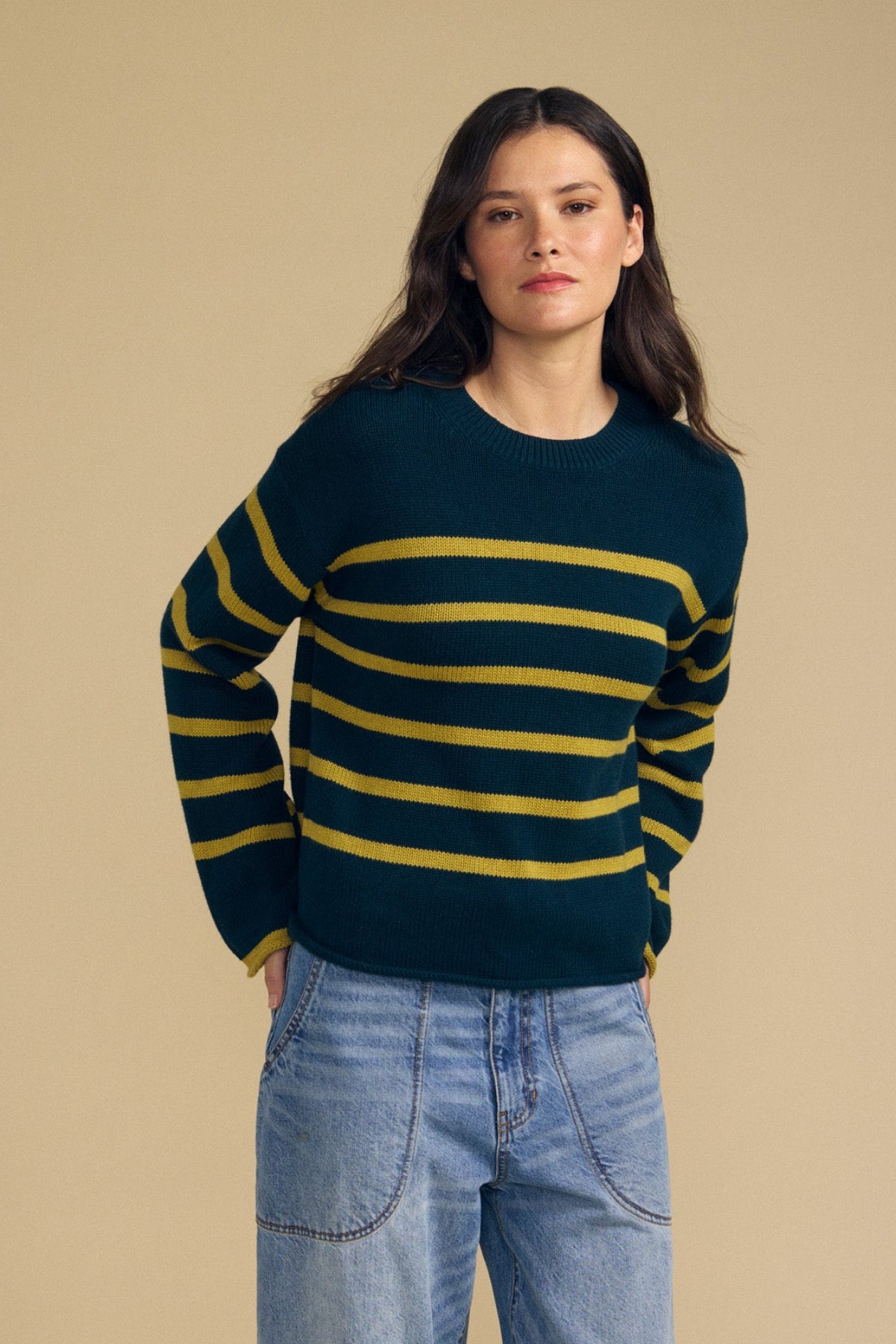 A person wearing the Velvet by Graham & Spencer LUCIA SWEATER—a relaxed-fit, dark green crew-neck sweater with yellow horizontal stripes—paired with blue jeans stands against a plain beige background.