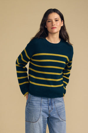 A person wearing the Velvet by Graham & Spencer LUCIA SWEATER—a relaxed-fit, dark green crew-neck sweater with yellow horizontal stripes—paired with blue jeans stands against a plain beige background.