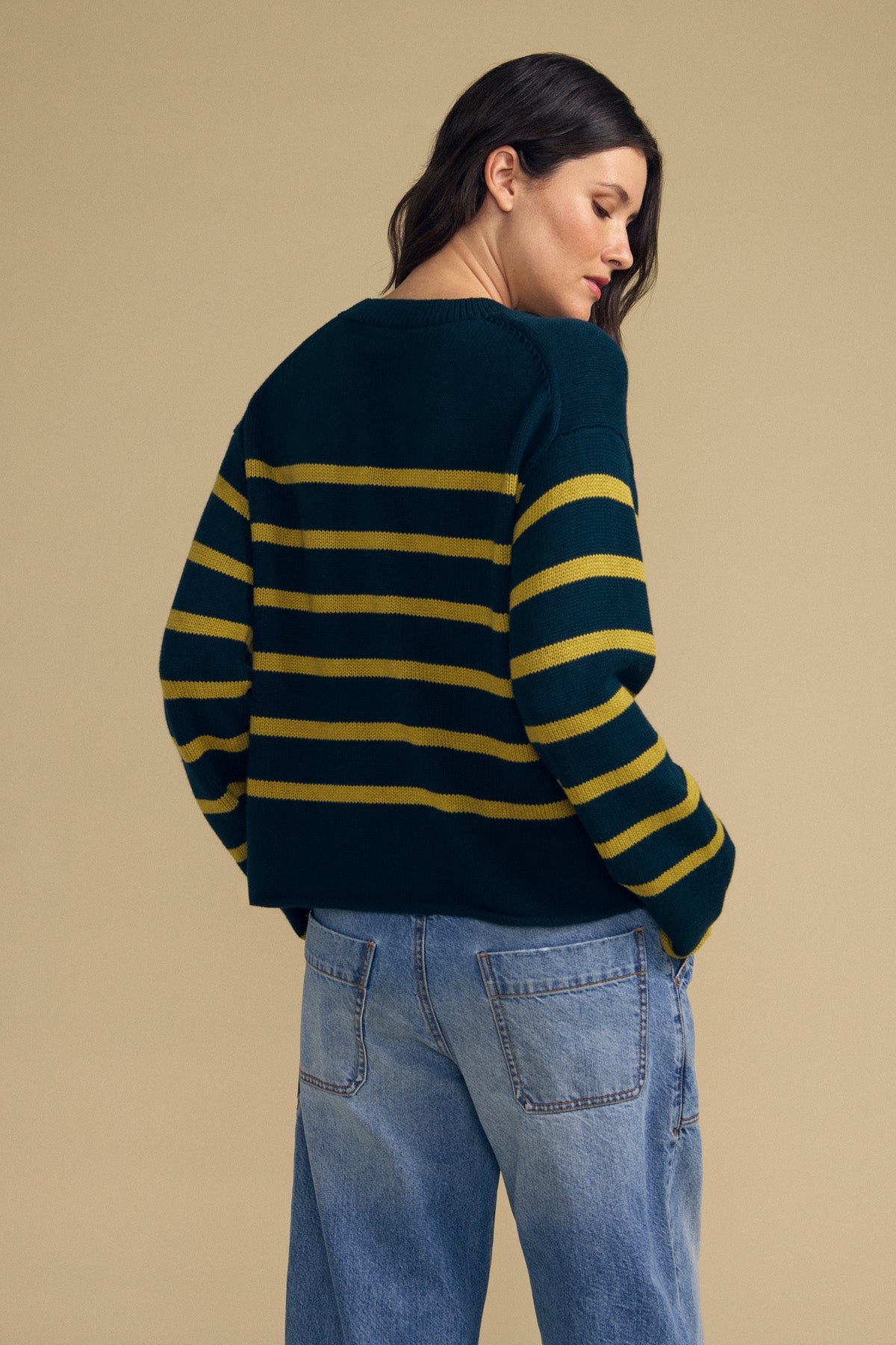 A person stands facing away against a plain beige background, wearing the LUCIA SWEATER by Velvet by Graham & Spencer—a dark green relaxed-fit sweater with yellow horizontal stripes—paired with light blue jeans.