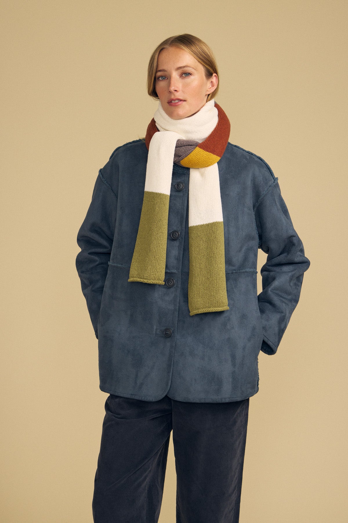 A person stands against a beige background with hands in their pockets, wearing the JADE SCARF by Velvet by Graham & Spencer, a blue button-up jacket, and dark pants.