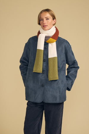 A person stands against a beige background with hands in their pockets, wearing the JADE SCARF by Velvet by Graham & Spencer, a blue button-up jacket, and dark pants.
