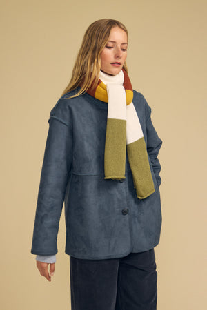 A woman wearing a blue oversized jacket and the Velvet by Graham & Spencer JADE SCARF—a striped, colorblocked accessory in mustard, white, and olive green—stands against a beige background.