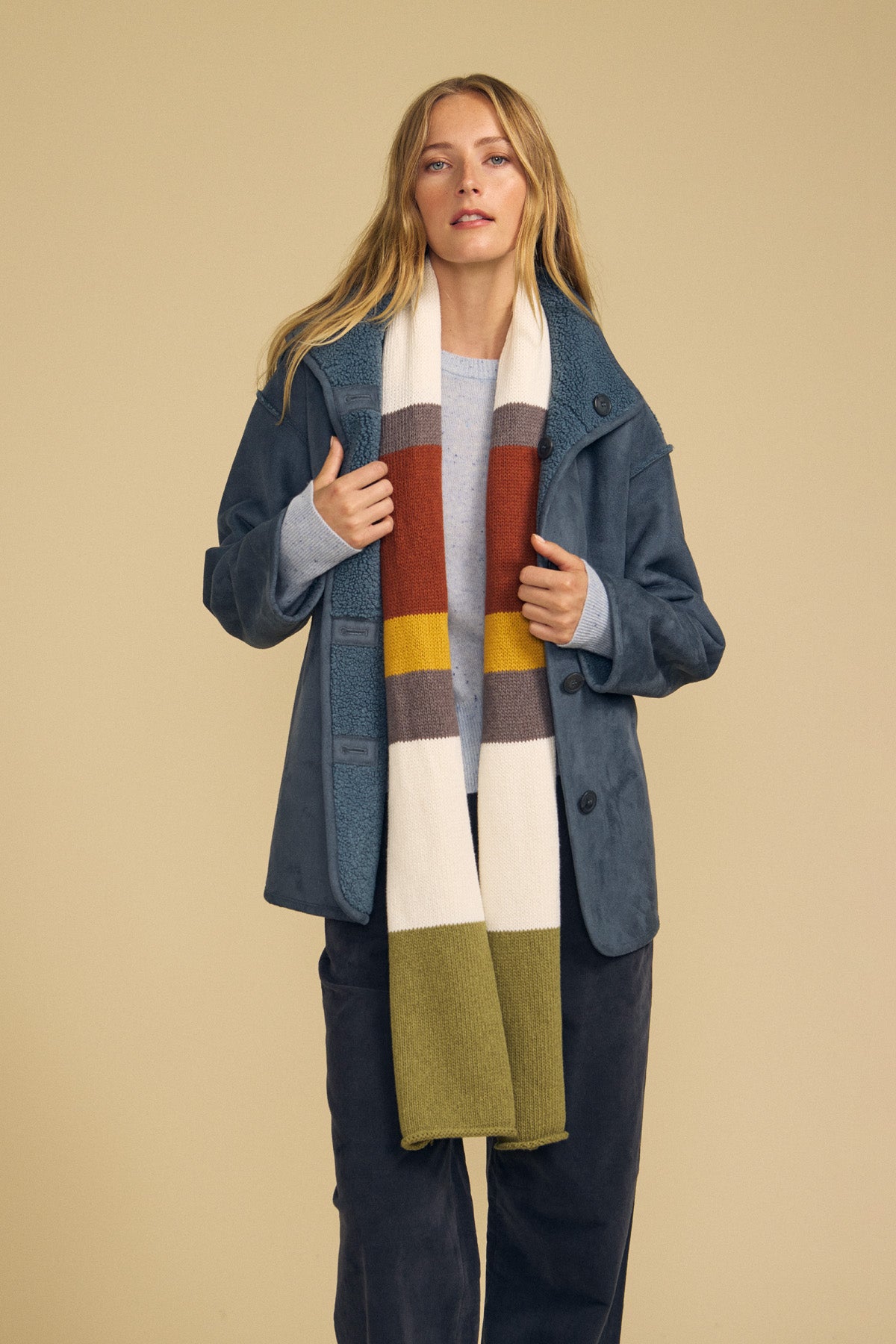 A woman stands against a beige background wearing a blue jacket, light blue sweater, and dark pants, styled with the Velvet by Graham & Spencer JADE SCARF—a long colorblocked accessory in cream, rust, gold, gray, and olive tones.