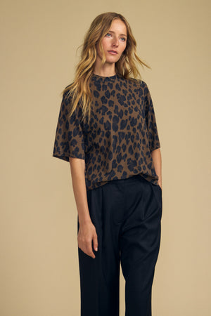 A woman stands against a plain beige background wearing the JANA TEE by Velvet by Graham & Spencer—a loose, short-sleeved women’s casual top with a brown and black leopard print on soft cotton jersey for all-day comfort, styled with high-waisted black pants.