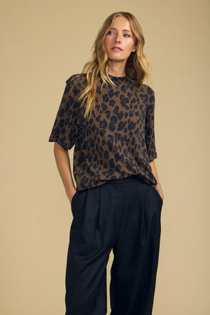 A woman stands against a beige background, one hand in her pocket, effortlessly showcasing the Velvet by Graham & Spencer JANA TEE—a loose leopard-printed women’s casual top crafted from soft cotton jersey—paired with black wide-leg trousers.