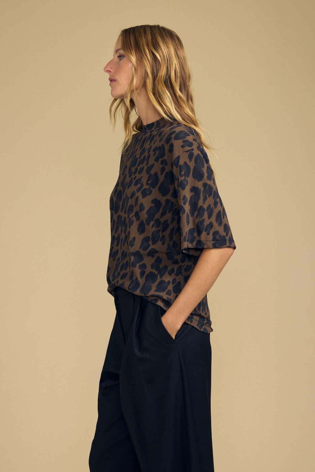 A woman in a side profile view wears the Velvet by Graham & Spencer JANA TEE, a loose leopard-printed cotton jersey top, paired with black pants and hands in pockets, standing against a beige background. This women’s casual tee offers everyday comfort with its soft cotton jersey fabric.