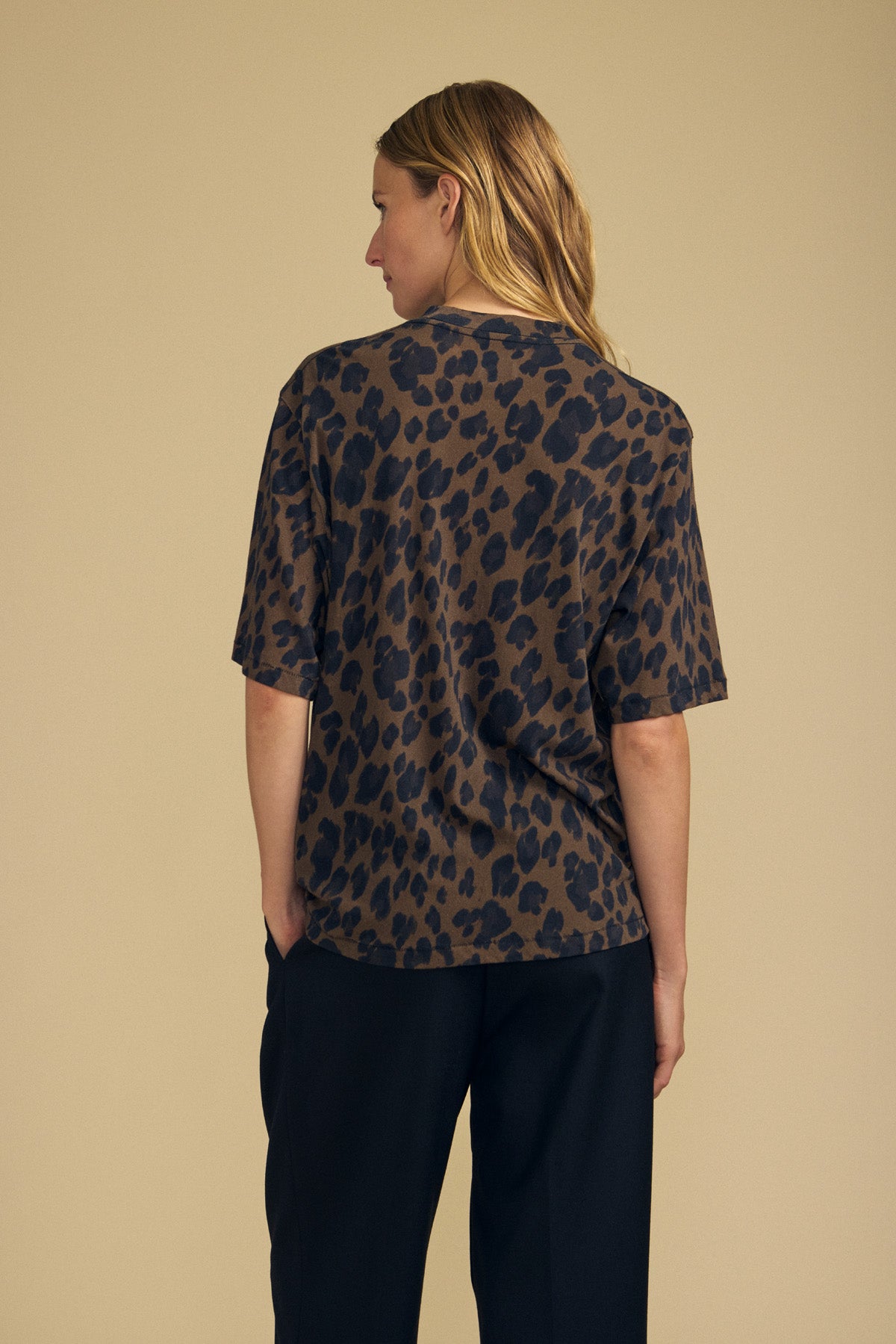 A person with long blonde hair stands facing away, wearing the JANA TEE by Velvet by Graham & Spencer—a loose-fitting, short-sleeve leopard print top paired with dark pants against a beige background. Crafted from comfortable cotton jersey, this women’s casual tee is perfect for everyday wear.