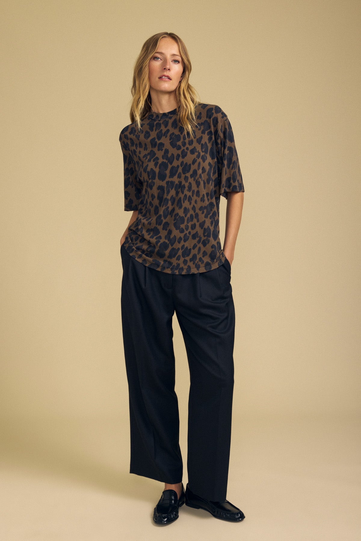 Against a beige background, a person stands with hands in pockets and a neutral expression, wearing the Velvet by Graham & Spencer JANA TEE—a loose cotton jersey leopard print top—paired with wide-leg black pants and black shoes.