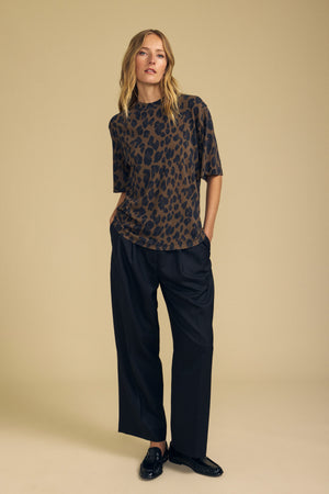 Against a beige background, a person stands with hands in pockets and a neutral expression, wearing the Velvet by Graham & Spencer JANA TEE—a loose cotton jersey leopard print top—paired with wide-leg black pants and black shoes.