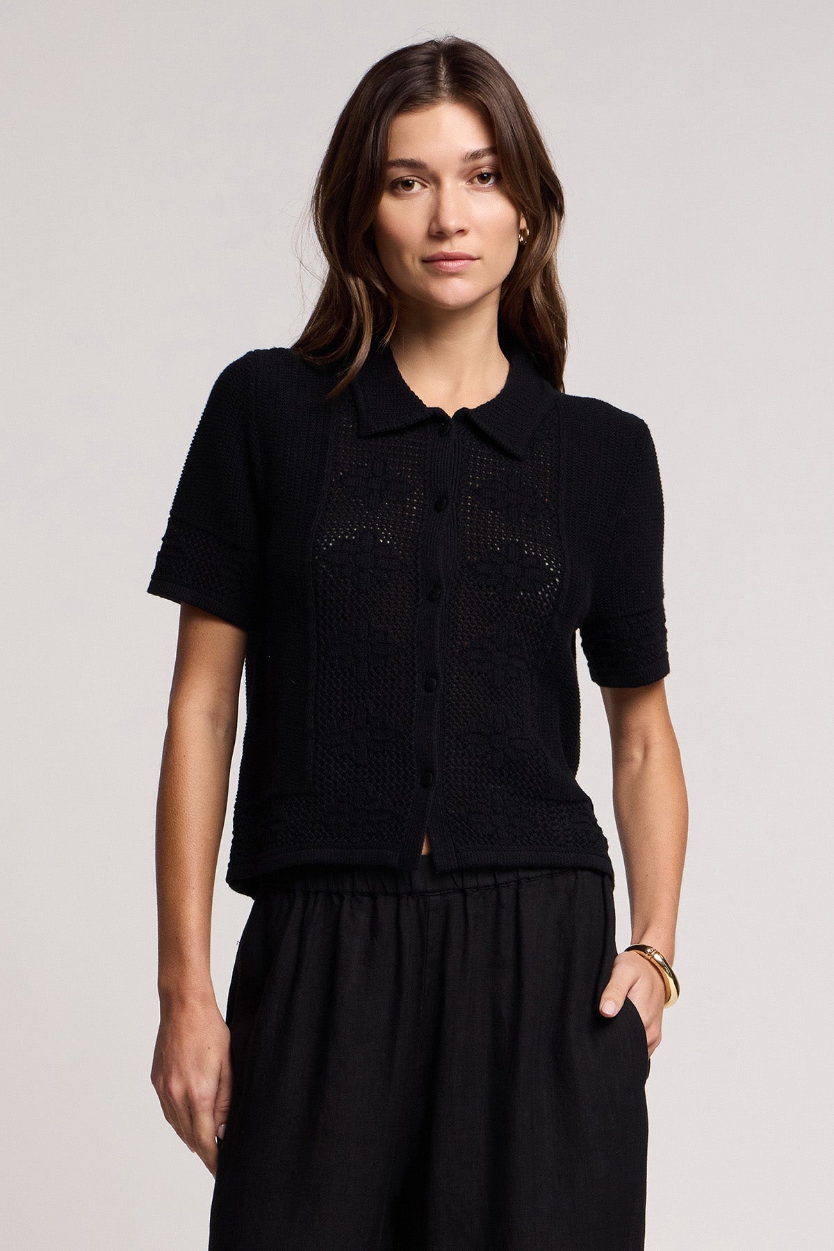 A woman wears the ROSEMOND CARDIGAN by Velvet by Graham & Spencer—a black short-sleeve button-up knit top with mesh stitching and a collar—paired with black pants, standing with one hand in her pocket against a plain background.-41372111896769