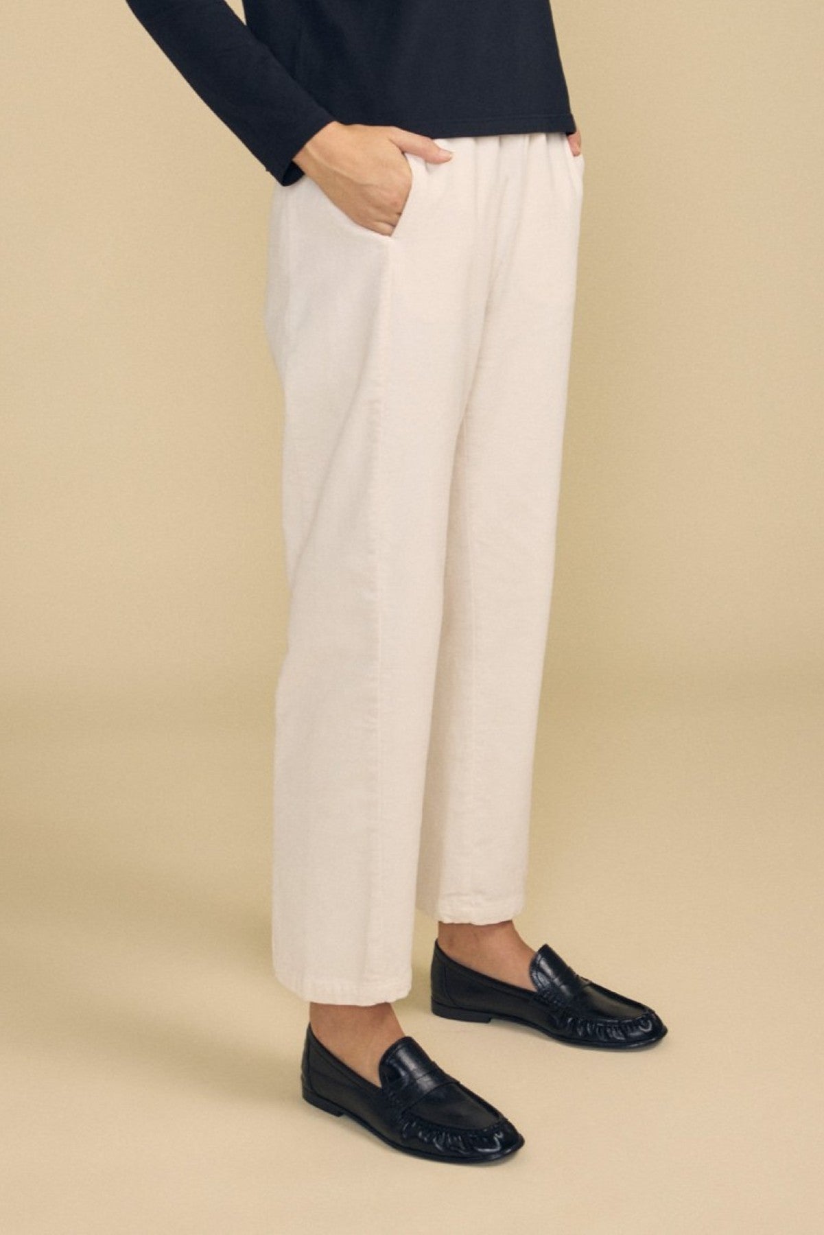 A person stands with hands in pockets, wearing the LANEY CORDUROY PANT by Velvet by Graham & Spencer—a light beige elastic waist pant—paired with a black long-sleeve top and black loafers, set against a plain beige background.