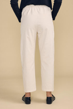 A person stands with their back to the camera, wearing the LANEY CORDUROY PANT by Velvet by Graham & Spencer in a cream pinwale corduroy with an elastic pull-on waist, paired with a black long-sleeve top and black shoes, set against a plain beige background.