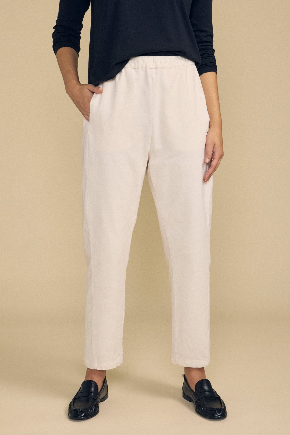 A person stands against a beige background wearing a black long-sleeve top, black loafers, and the LANEY CORDUROY PANT by Velvet by Graham & Spencer—cream-colored cropped pants with an elastic waist crafted from pinwale corduroy.