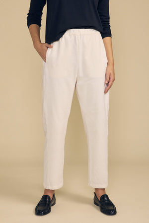 A person stands against a beige background wearing a black long-sleeve top, black loafers, and the LANEY CORDUROY PANT by Velvet by Graham & Spencer—cream-colored cropped pants with an elastic waist crafted from pinwale corduroy.