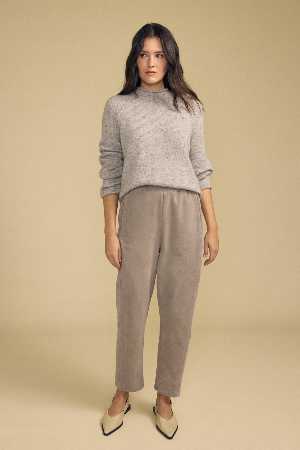 A woman stands against a beige background, dressed in a light gray sweater and the LANEY CORDUROY PANT by Velvet by Graham & Spencer—a taupe pinwale corduroy pant with a cropped length and an elastic pull-on waist—paired with cream-colored heels; her expression is neutral and her posture relaxed.