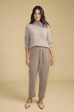 A woman stands against a beige background, dressed in a light gray sweater and the LANEY CORDUROY PANT by Velvet by Graham & Spencer—a taupe pinwale corduroy pant with a cropped length and an elastic pull-on waist—paired with cream-colored heels; her expression is neutral and her posture relaxed.