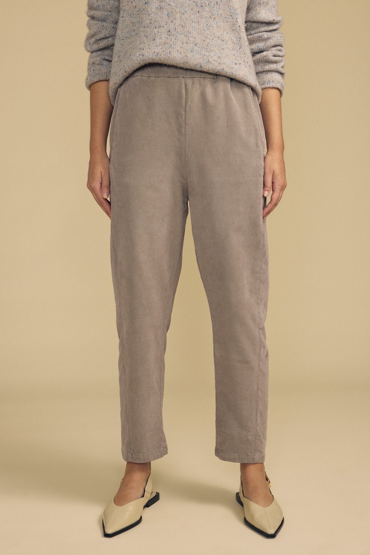 A person stands in front of a plain beige background wearing a light gray sweater, Velvet by Graham & Spencer’s LANEY CORDUROY PANT in taupe with a comfortable pull-on waist, and cream pointed-toe flats.