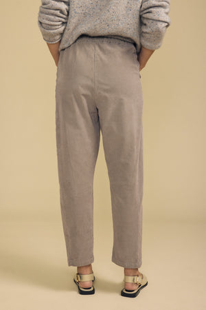 A person stands with their back to the camera against a plain beige background, wearing a speckled sweater, cream sandals, and the LANEY CORDUROY PANT by Velvet by Graham & Spencer—a loose-fitting, beige pull-on style.