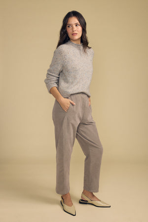 A woman stands with her hands in her pockets, wearing a light gray sweater and the LANEY CORDUROY PANT by Velvet by Graham & Spencer—a beige pinwale corduroy cropped pant with an elastic waist—paired with light-colored flats, set against a plain beige background.