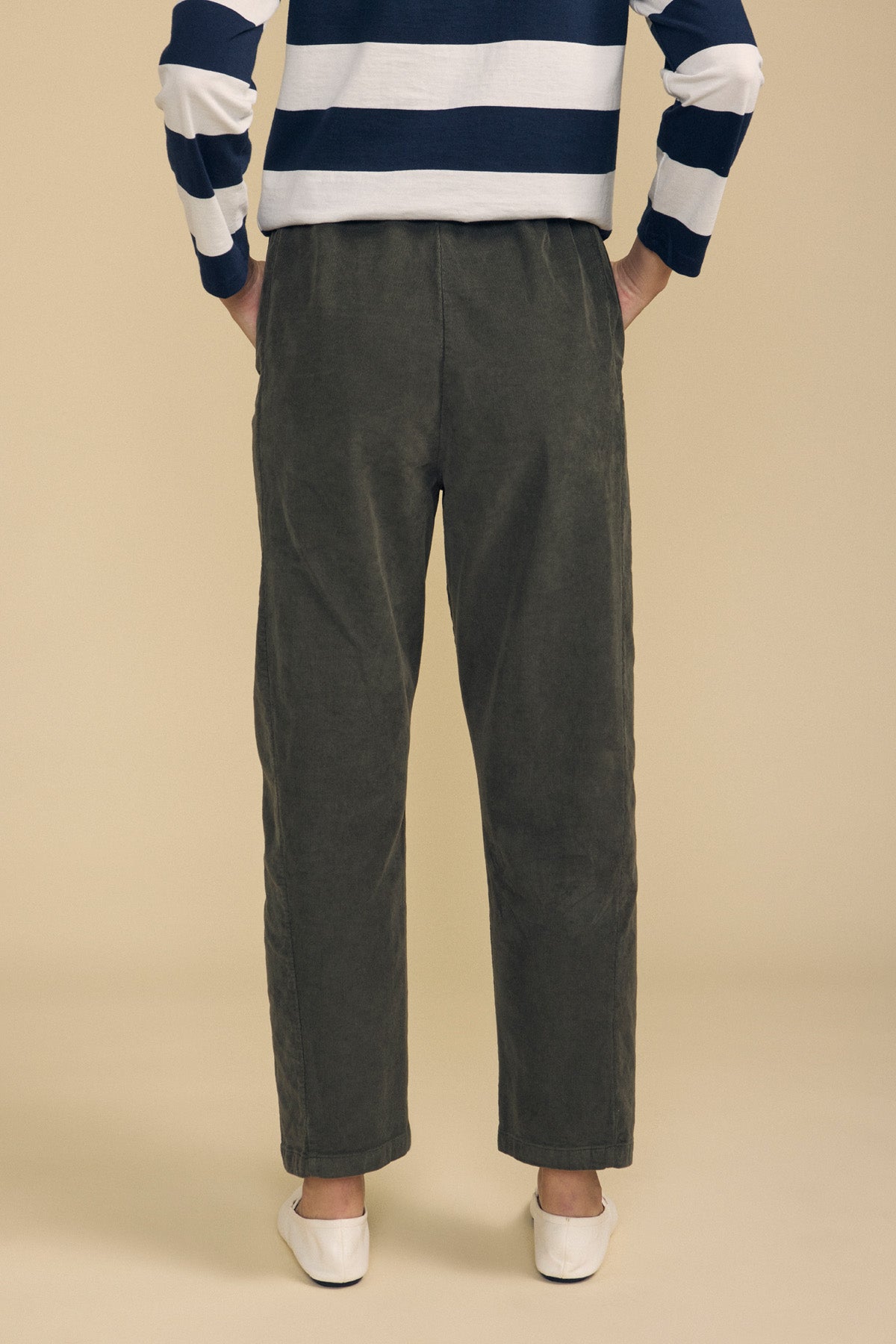 A person stands with hands on hips, facing away from the camera against a beige background, wearing Velvet by Graham & Spencer’s LANEY CORDUROY PANT in olive green with a pull-on waist, paired with a navy and white striped long-sleeve shirt and white flat shoes.