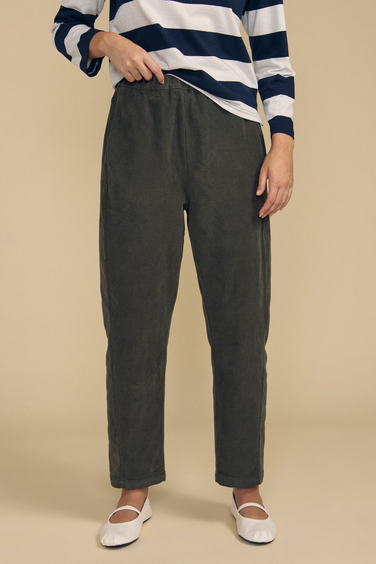 Wearing the LANEY CORDUROY PANT by Velvet by Graham & Spencer in dark gray with a pull-on waist, a person pairs them with a navy and white striped long-sleeve shirt and white ballet flats while standing against a beige background.-41860470177985