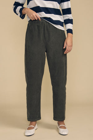 Wearing the LANEY CORDUROY PANT by Velvet by Graham & Spencer in dark gray with a pull-on waist, a person pairs them with a navy and white striped long-sleeve shirt and white ballet flats while standing against a beige background.