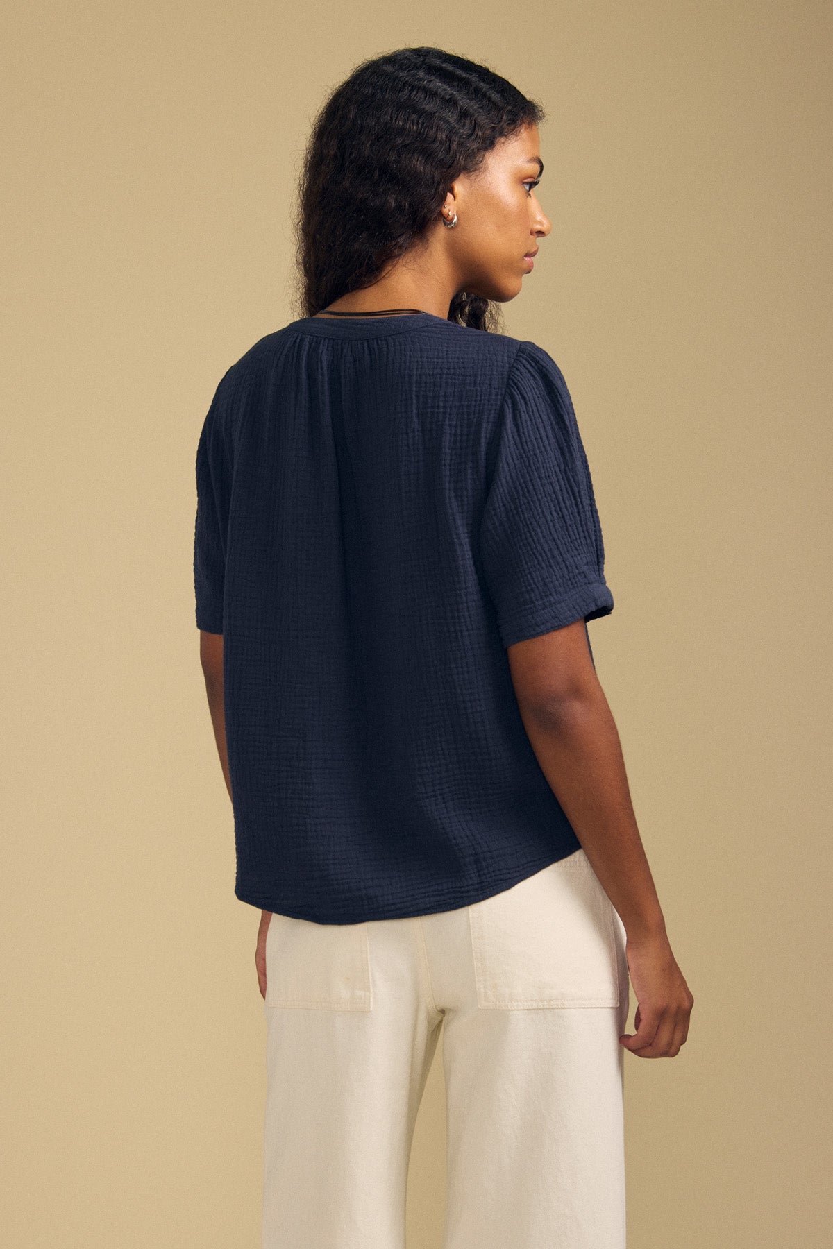 A person stands against a beige background, seen from behind and turned slightly to the side, wearing the Velvet by Graham & Spencer DEANN TOP—a navy blue, relaxed-fit cotton gauze short-sleeve top—paired with off-white pants.-41758773313729