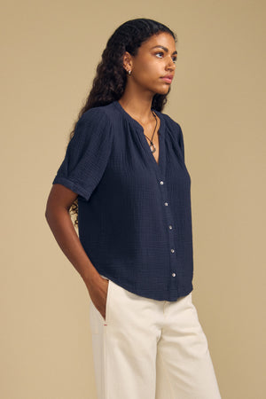 A person with long curly hair wears the DEANN TOP by Velvet by Graham & Spencer, a relaxed-fit navy cotton gauze button-up, paired with off-white pants and standing against a plain beige background with hands in their pockets.
