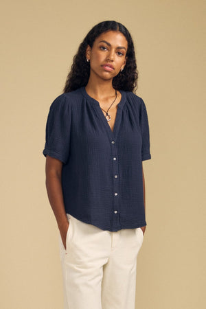 A woman stands against a beige background, wearing the DEANN TOP by Velvet by Graham & Spencer—a relaxed-fit navy short-sleeve button-up shirt with a subtle v-neckline—paired with cream trousers and black flats. Her hands are in her pockets, and she has long curly hair.