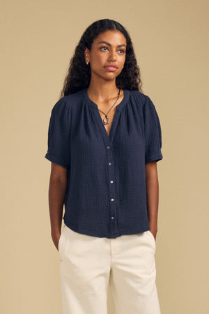 A woman with curly hair stands against a beige background wearing the Velvet by Graham & Spencer DEANN TOP, a navy, relaxed-fit piece featuring short sleeves and a subtle v-neckline, paired with white wide-leg pants, black shoes, and jewelry. Her hands are in her pockets.
