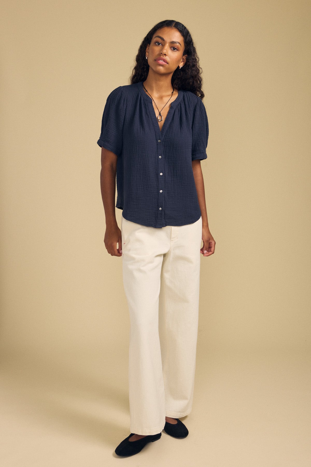 A person stands against a beige background wearing the DEANN TOP by Velvet by Graham & Spencer—a relaxed-fit black blouse with a v-neckline—paired with cream wide-leg pants and black shoes.-41758773051585