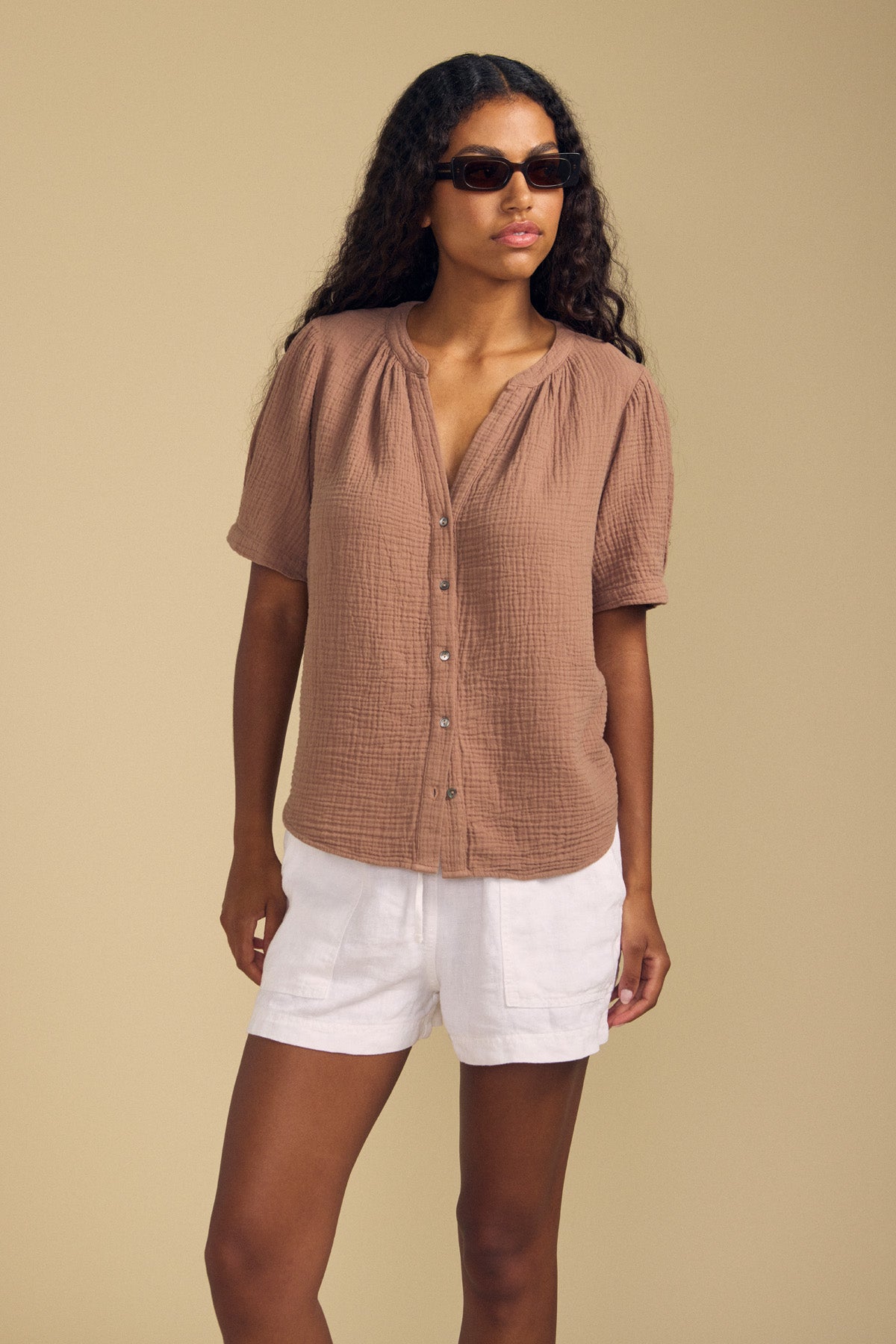 A woman wearing sunglasses and the DEANN TOP by Velvet by Graham & Spencer—a relaxed-fit cotton gauze top featuring a v-neckline and short sleeves—paired with white shorts, stands against a plain beige background.