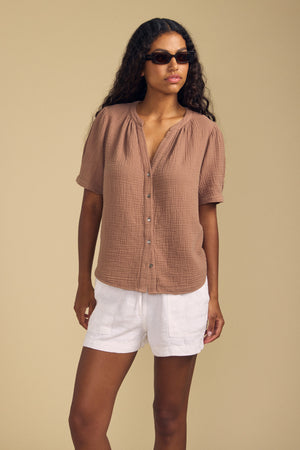 A woman wearing sunglasses and the DEANN TOP by Velvet by Graham & Spencer—a relaxed-fit cotton gauze top featuring a v-neckline and short sleeves—paired with white shorts, stands against a plain beige background.