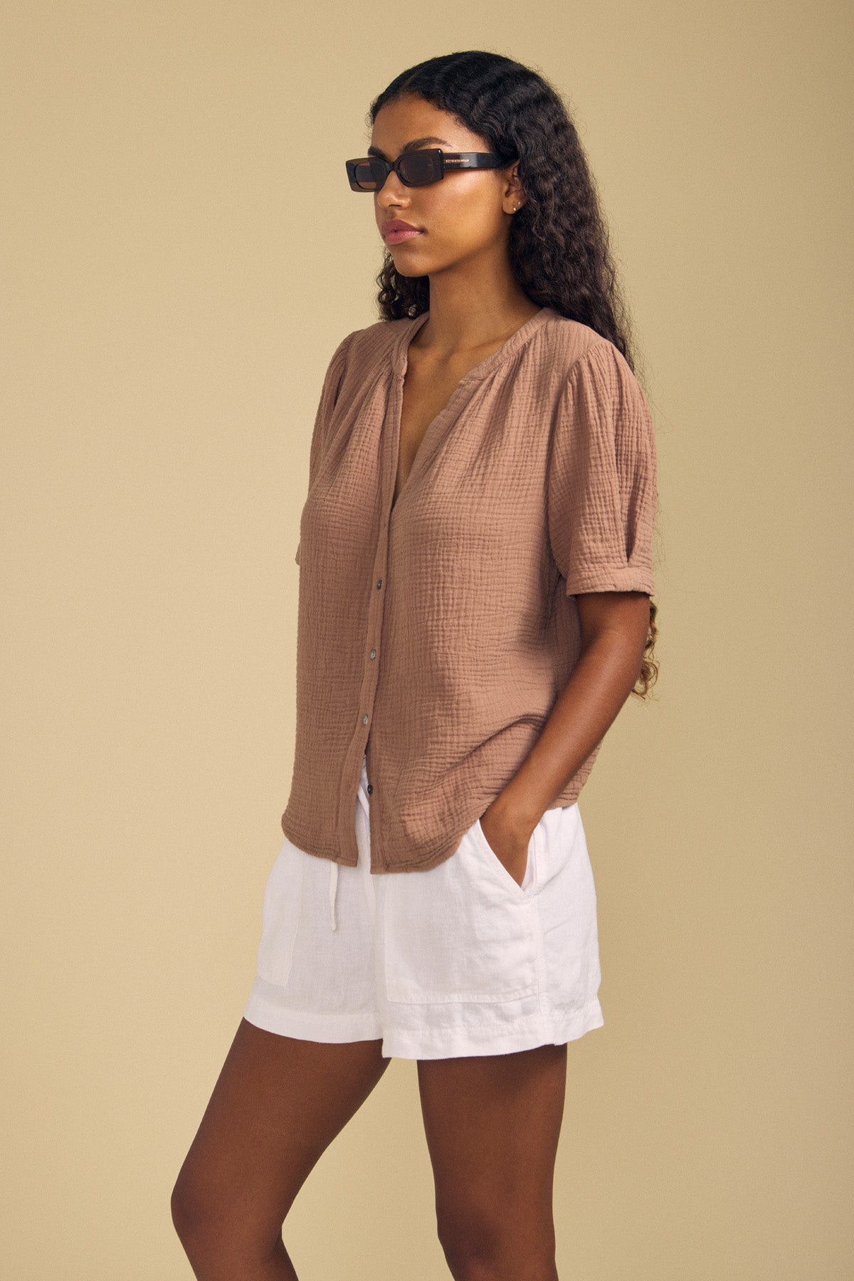A woman wearing the DEANN TOP by Velvet by Graham & Spencer—a relaxed-fit brown button-up shirt with a subtle v-neckline—pairs it with white shorts and sunglasses, standing against a beige background with her hands in her pockets.