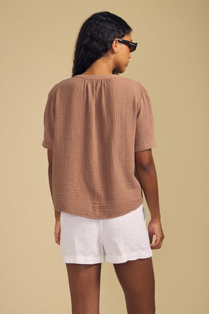 A person with long dark hair, wearing sunglasses and the DEANN TOP by Velvet by Graham & Spencer—a textured brown cotton gauze v-neck blouse—paired with white shorts, stands facing away from the camera against a plain beige background.