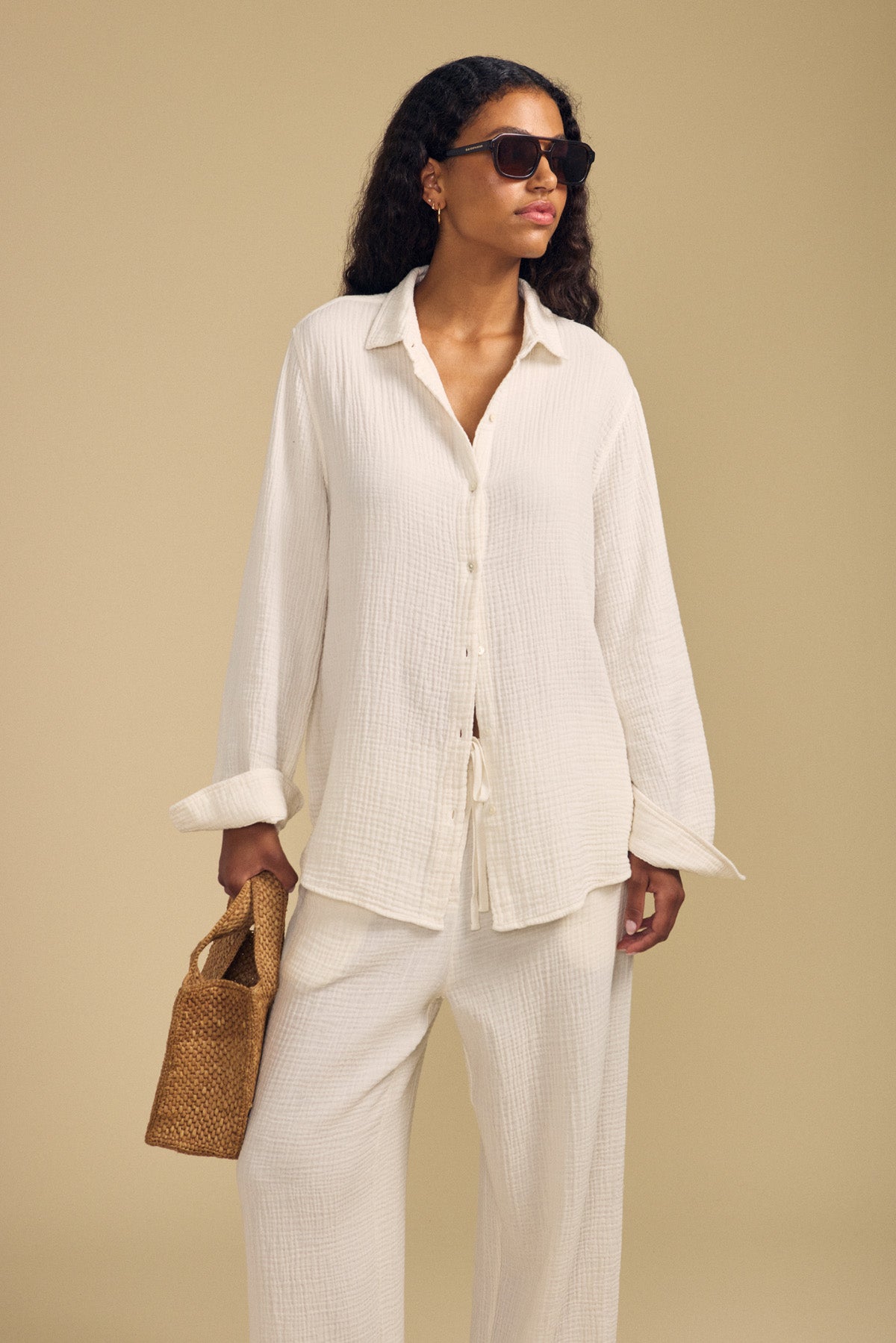 A woman stands against a neutral background wearing the DENISE SHIRT by Velvet by Graham & Spencer—a loose, cream-colored cotton gauze button-up—paired with matching pants, a woven tote bag, and dark sunglasses.